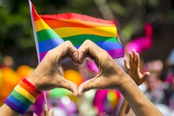 A photo of a hand heart in the foreground and colorful pride flags in the background.