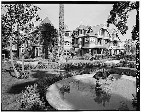A black and white photo of the Winchester House, a large Victorian Era mansion. Trees and a fountain are in the foreground.