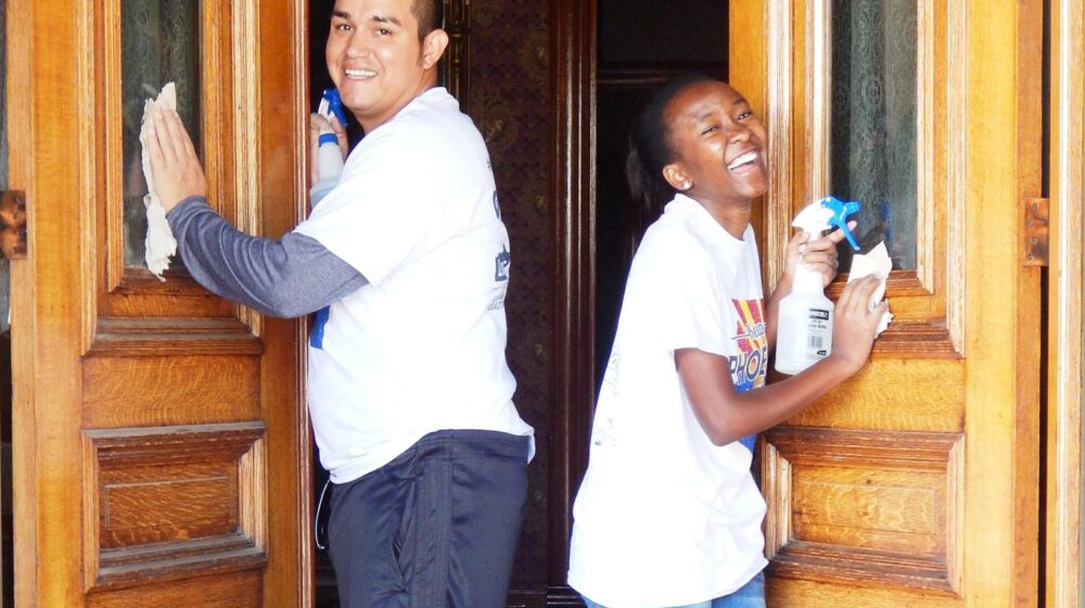 Two The Square PHX volunteers smile and laugh as they clean the windows in the front doors of Rosson House.