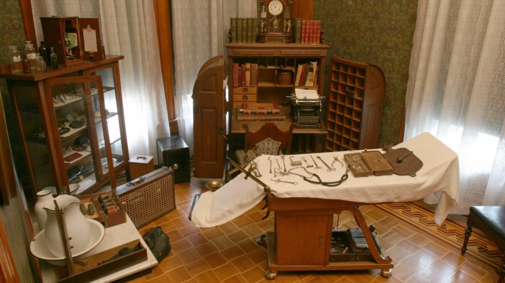 The doctor’s office at Rosson House, with dark green wallpaper, parquet wood flooring, a cabinet desk, an examining table and many other antique medical instruments.