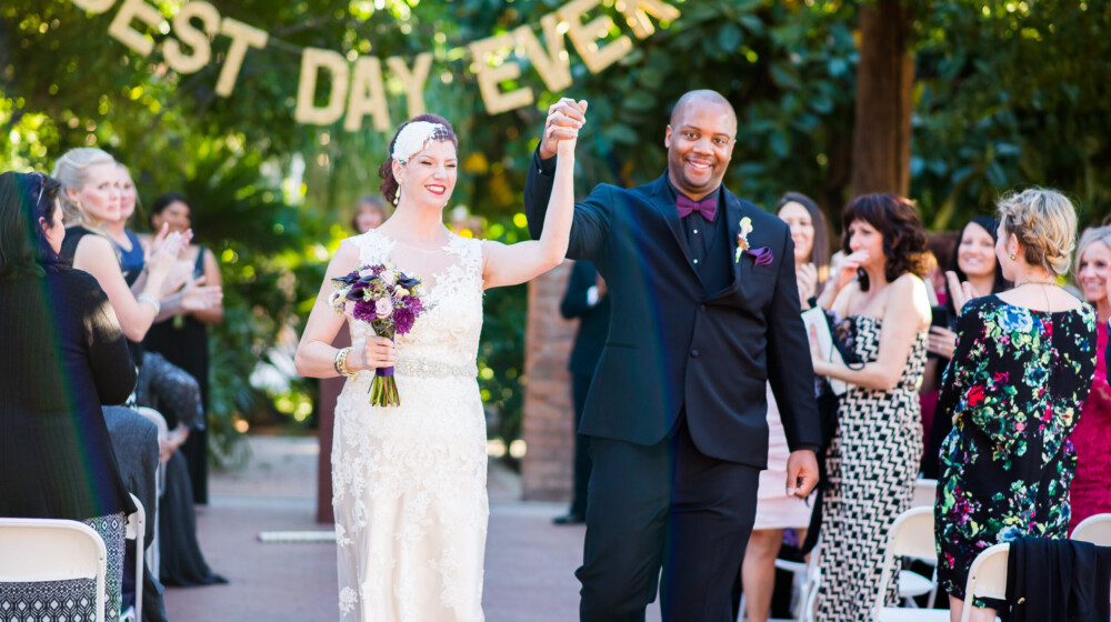 A bride and groom at their wedding at The Square PHX, holding hands and lifting them in celebration, with the words 