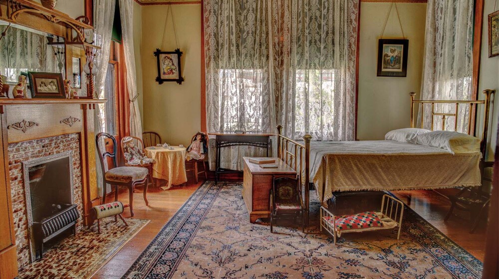 The north bedroom at Rosson House, with light green wallpaper, a fireplace, a brass double bed, and several antique toys.