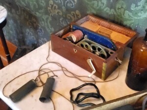 A picture of our electrotherapy machine from approximately 1870. The machine comes in a medium-sized wooden box, and has gears and fabric inside to create static electricity that builds up and is transferred to the hand of the person holding the metal handles that are attached to the box with wires.