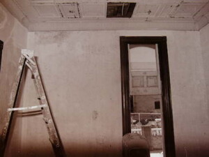 A picture of the Rosson House formal parlor during restoration. The walls have been stripped of wallpaper and work is being done on the pressed metal ceiling. The vertical pocket door is half raised so the Burgess Carriage House can be seen past work being done on the front porch.