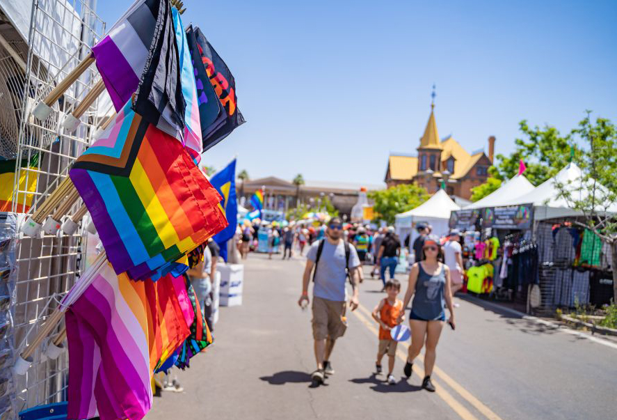 A picture of the Rainbows Festival at The Square PHX, with festival tents and booths lining the street, people enjoying themselves, beautiful and colorful pride flags, and Rosson House in the background.