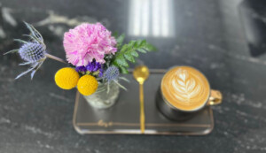 A picture of a tasty looking cup of coffee with a heart shaped latte art in the foam on top, next to bright and beautiful looking flowers.