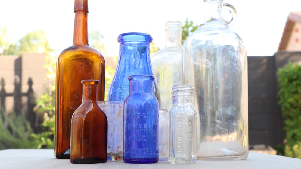 A picture of nine bottles or glass containers, all of different shapes and sizes, from the The Square PHX museum collection. There are two brown bottles, two cobalt blue bottles, and the remainder are clear glass.