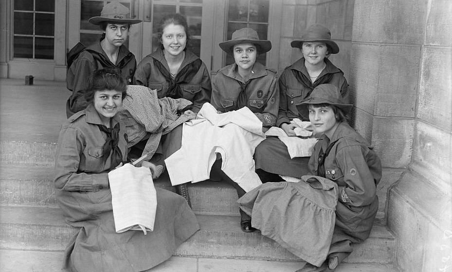 A black and white picture, taken in 1918, of six Girl Scouts in their uniforms, sitting on steps and sewing.