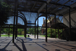 A picture from inside the Lath Pavilion at The Square PHX, looking out at the Rosson House Museum and Visitor Center. Verdant green grass and trees and a perfect, cloudless blue sky can be seen beyond.