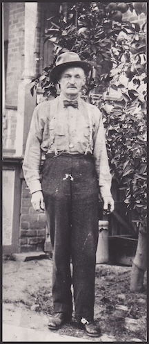 An old sepia photo of an older man with slacks and a button up shirt and hat, standing next to the corner of a brick building in front of a citrus tree.