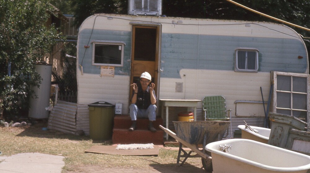 A picture of an older man, sitting in the doorway of a trailer in a construction area, with construction debris in the foreground.