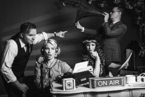 A present day black and white photo of people acting during a stage play of War of the Worlds, showing a radio program desk and four people.