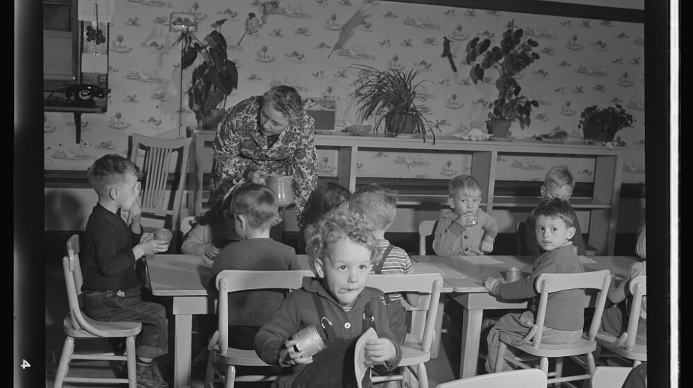 A black and white photo from 1943 showing a daycare facility with children eating lunch, and an adult helping.